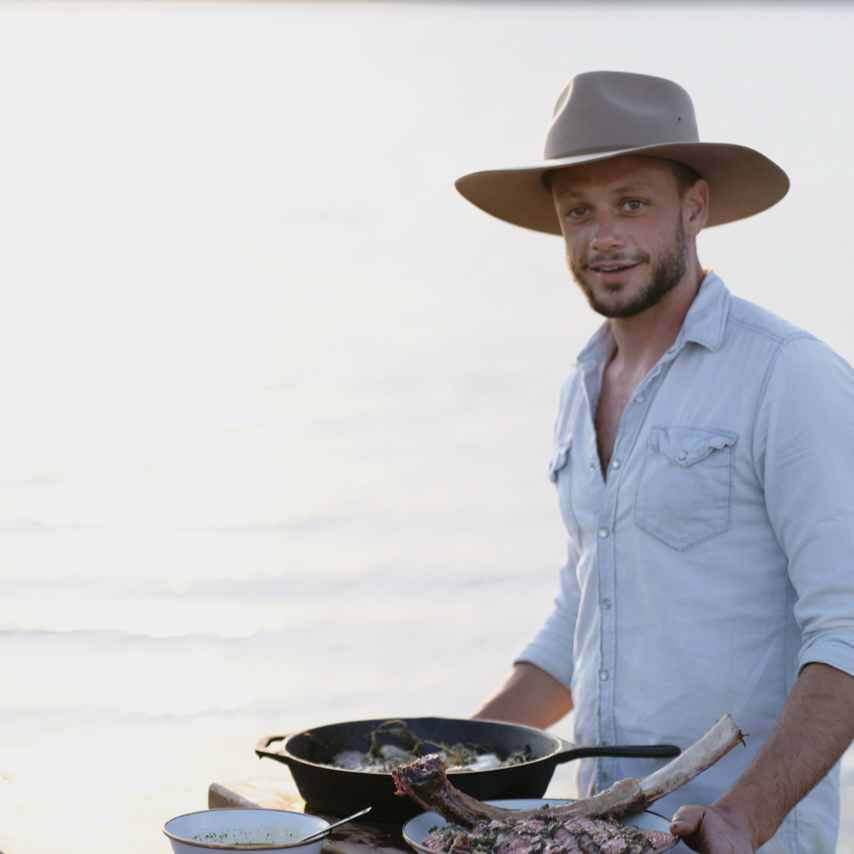 Man in a hat preparing food by a lake at sunset