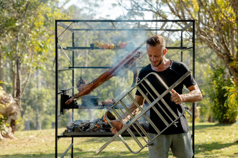 Man using a portable barbecue grill outdoors with trees in the background. A man tending to food on a large Portico open fire grill