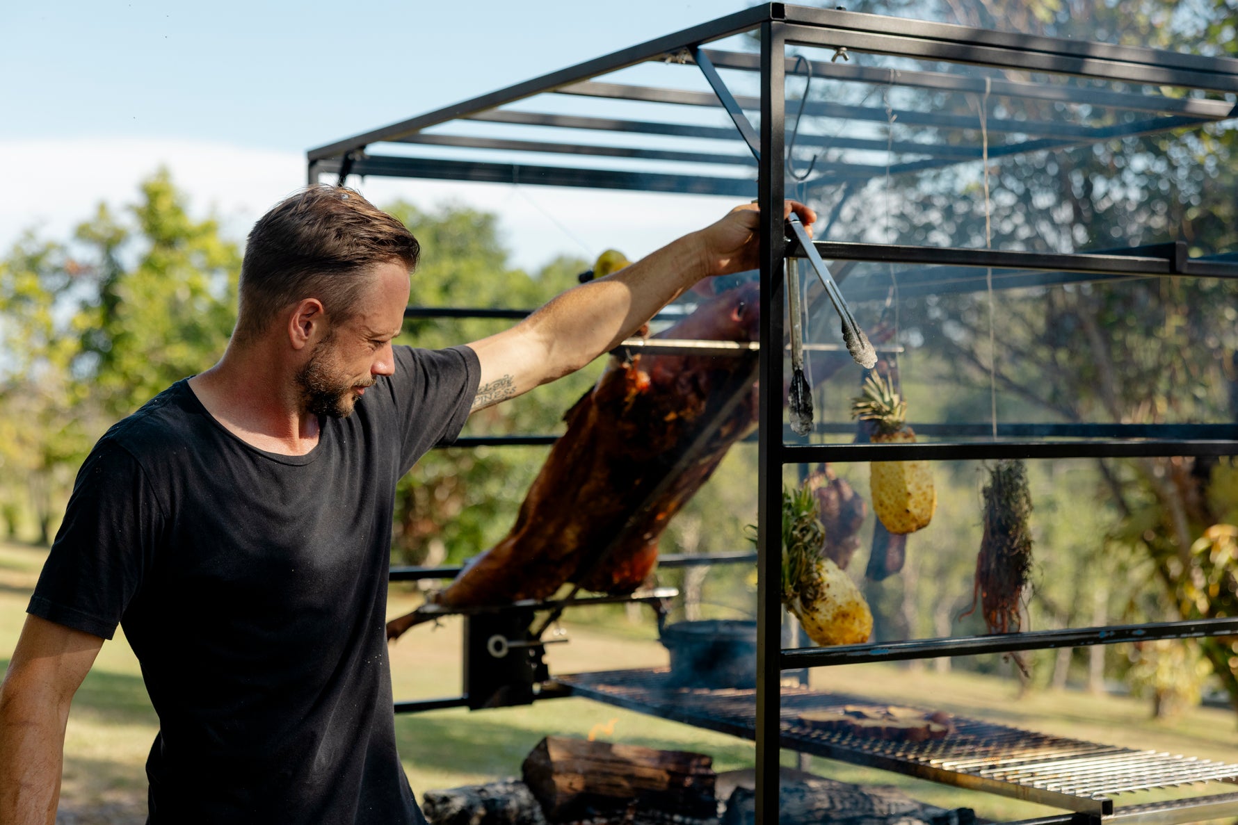 Man cooking food on a grill outdoors with trees in the background