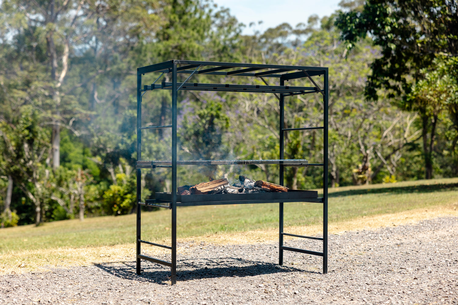 Outdoor barbecue grill with smoking meat on a gravel area with trees in the background. Portico Small Asado BBQ close-up of premium Australian-made construction