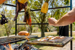 Person grilling food on an outdoor barbecue with trees in the background. Portico Large Churrasco Bundle detail showing premium grilling equipment