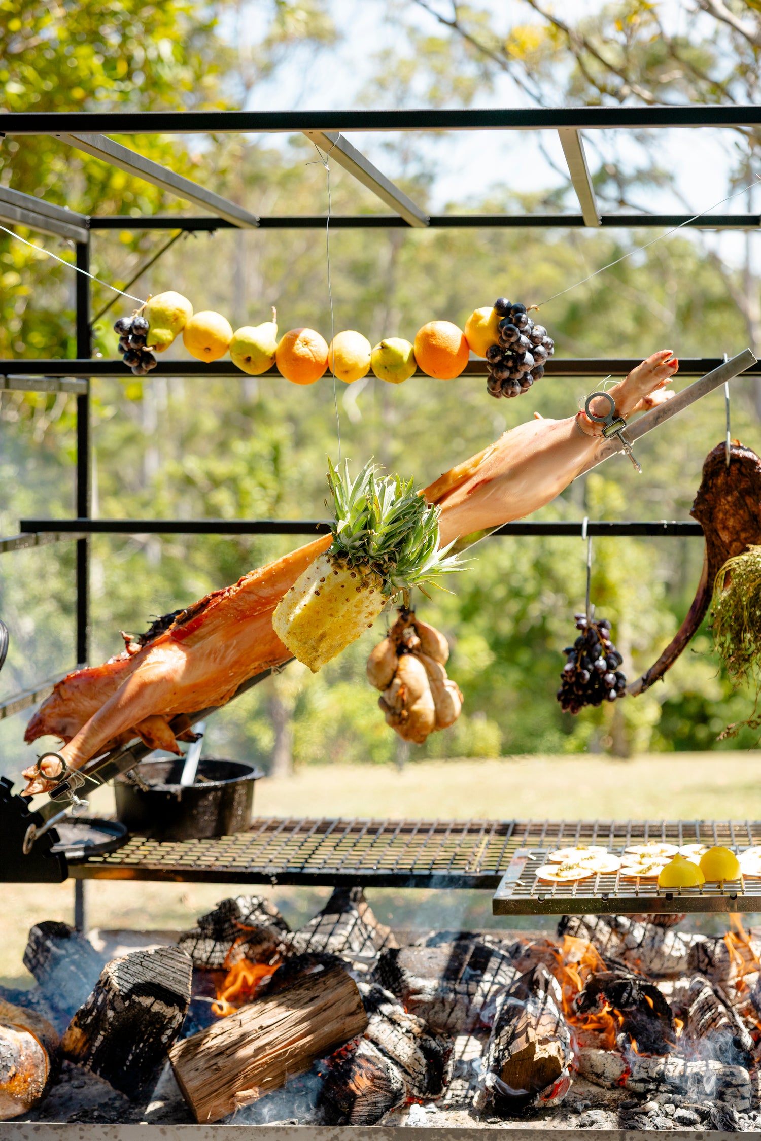 Hanging meats and vegetables over an open fire in a rustic setting with greenery. Chef Charlie Bundle showing Portico Large BBQ with all cooking accessories