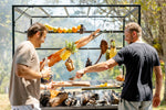Two men grilling food outdoors with a  Portico Argentinian Stylle BBQ from Terra Grill and various food items.