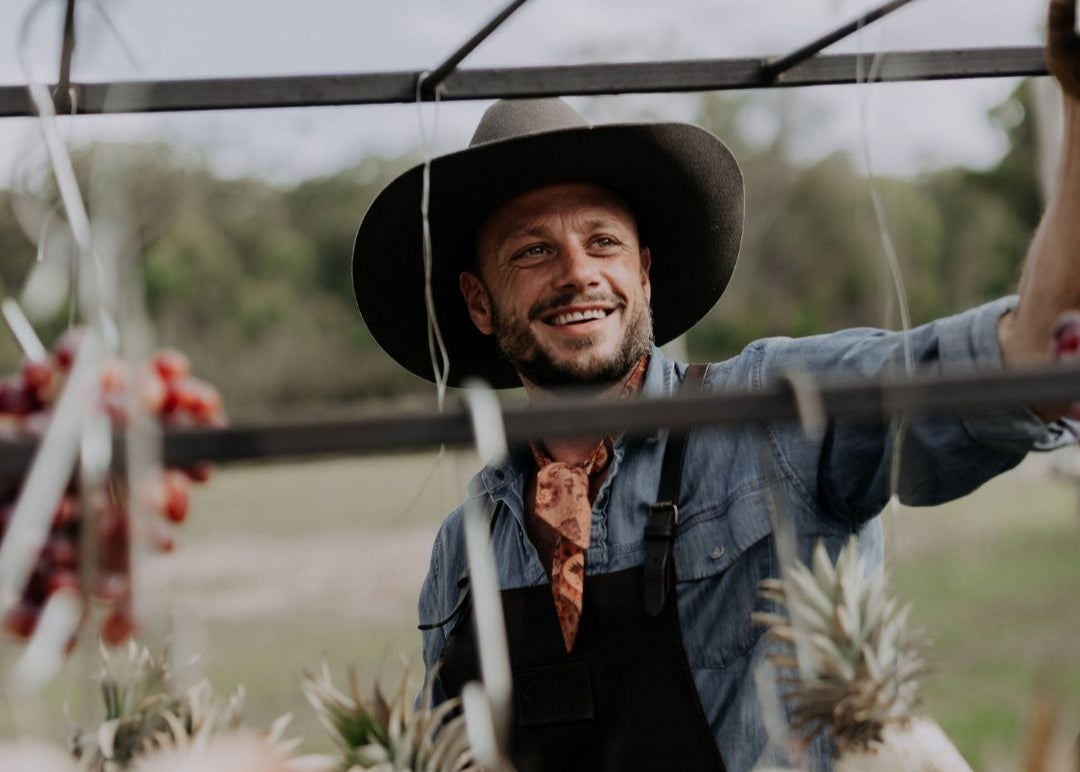 Man wearing a cowboy hat and overalls standing outdoors with plants and hanging fruit in the foreground. Cooking on the Argentinian style bbq from Terra Grills, thePortico